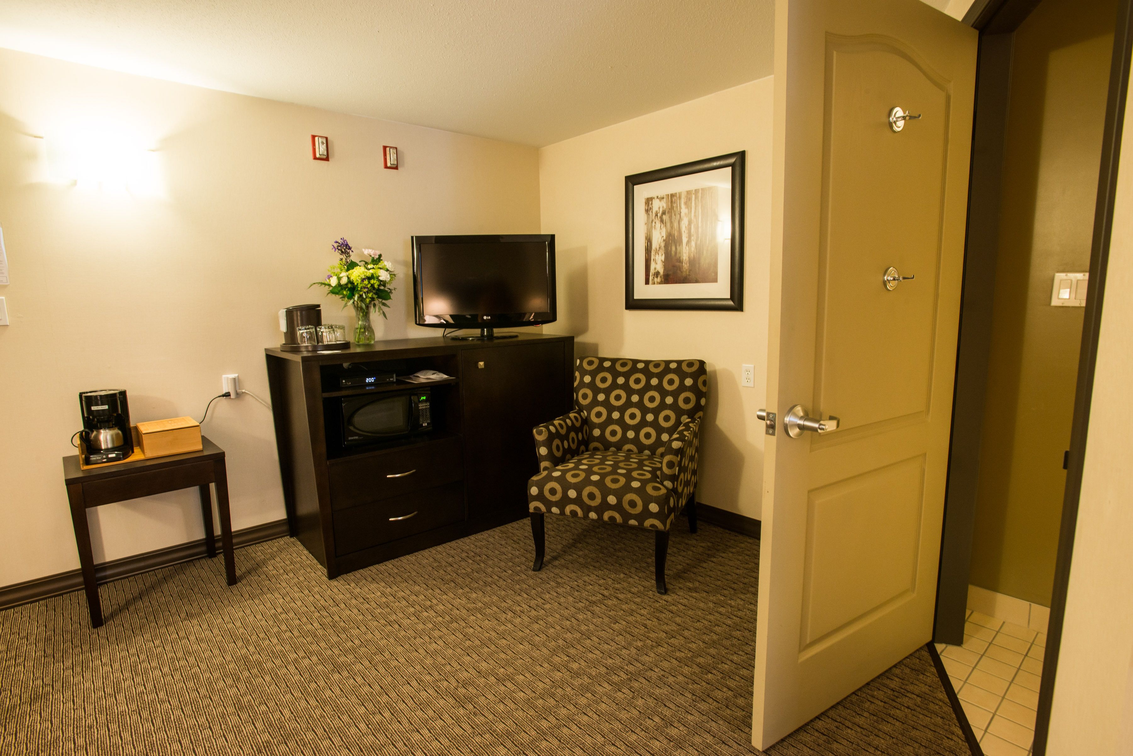 A hotel room with a television, a coffee maker, a vase with flowers, a patterned armchair, and a door.