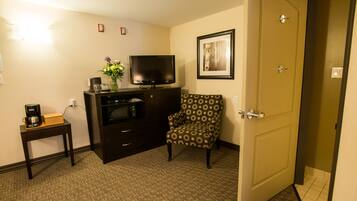 A hotel room with a television, a coffee maker, a vase with flowers, a patterned armchair, and a door.