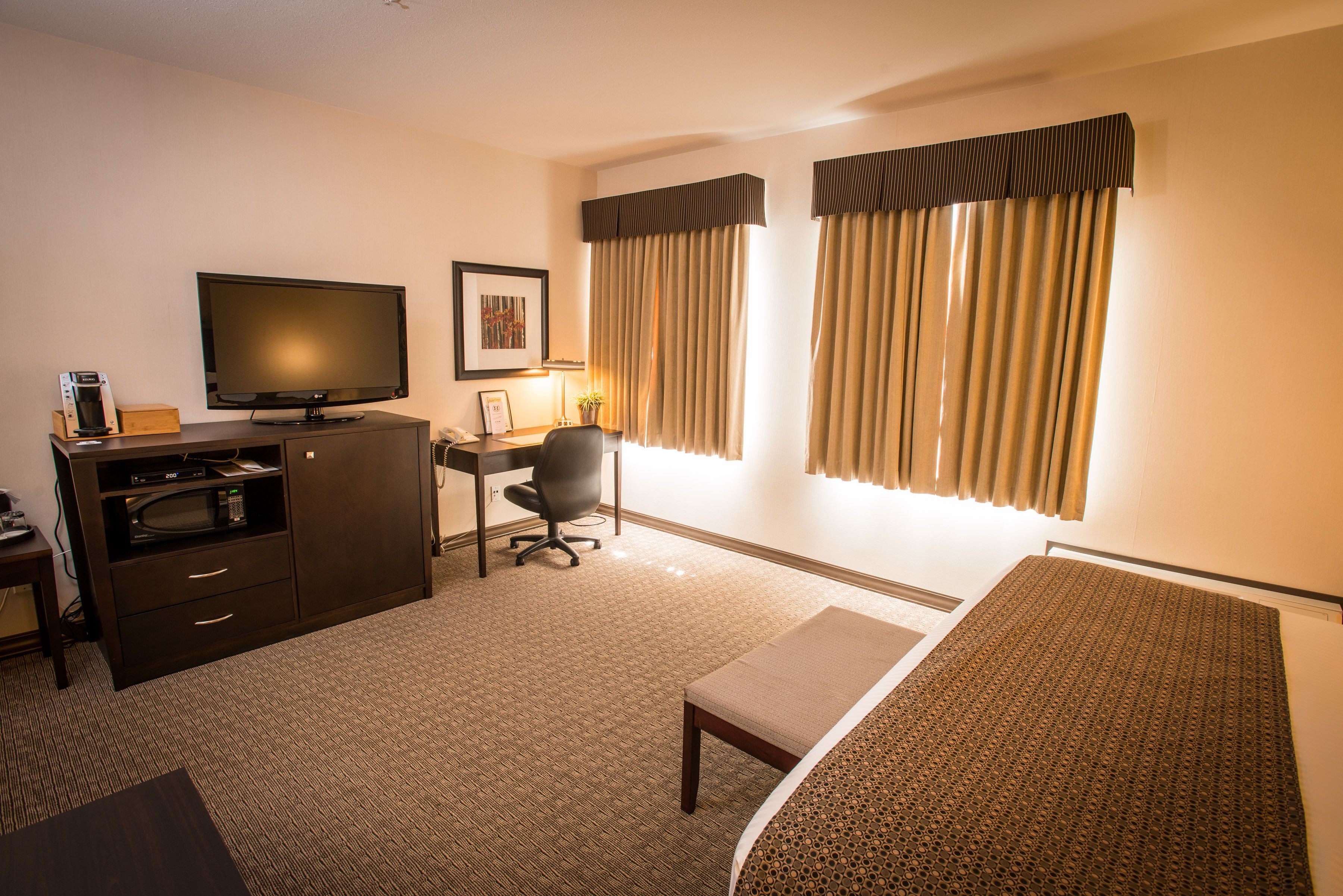 A hotel room with a bed, a desk with a chair, a television, a minibar, and a window with curtains.
