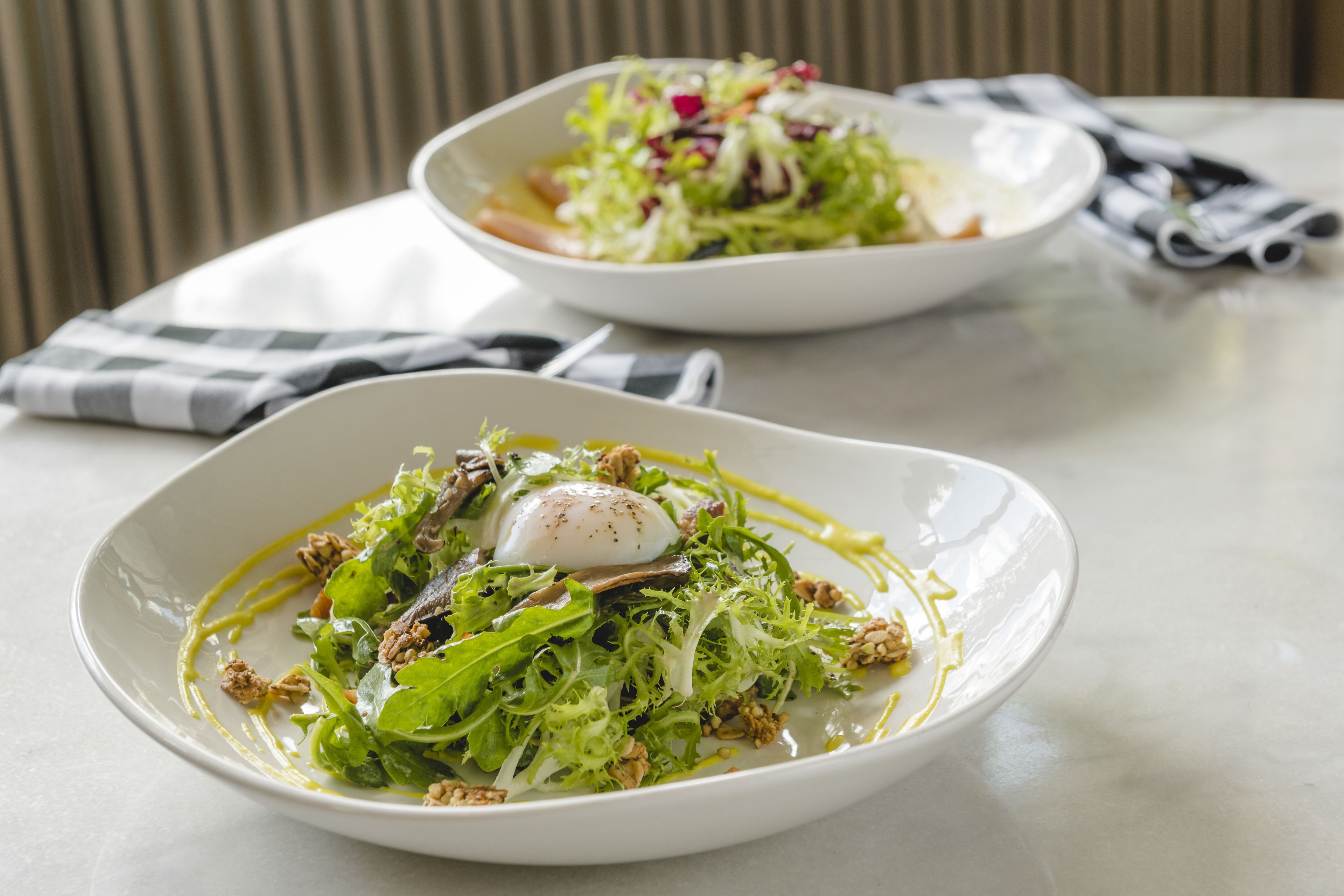 Two white plates of salad with a poached egg on a white table.