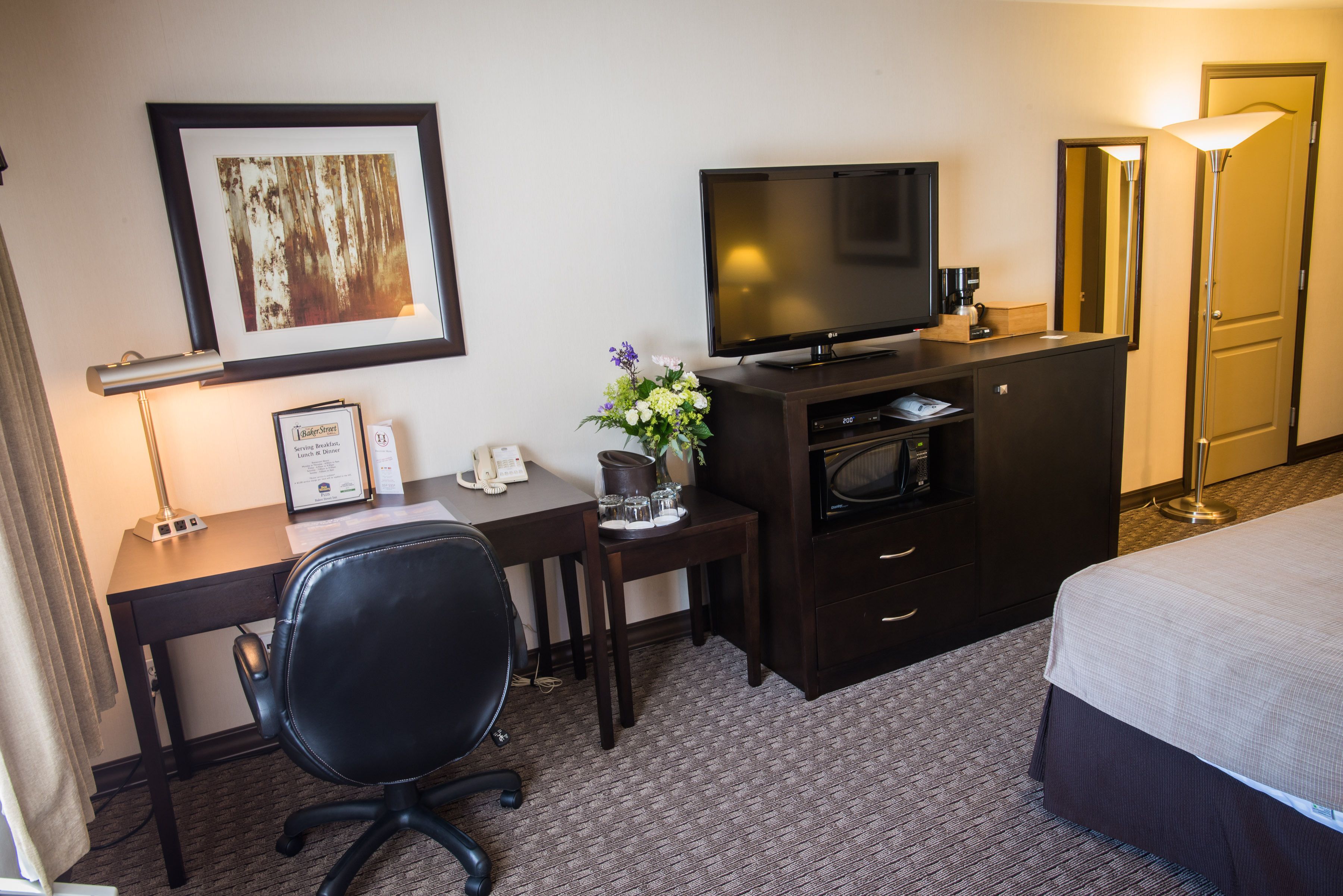 A hotel room with a bed, a desk with a chair, a television, a dresser, a framed picture on the wall, and a vase with flowers.