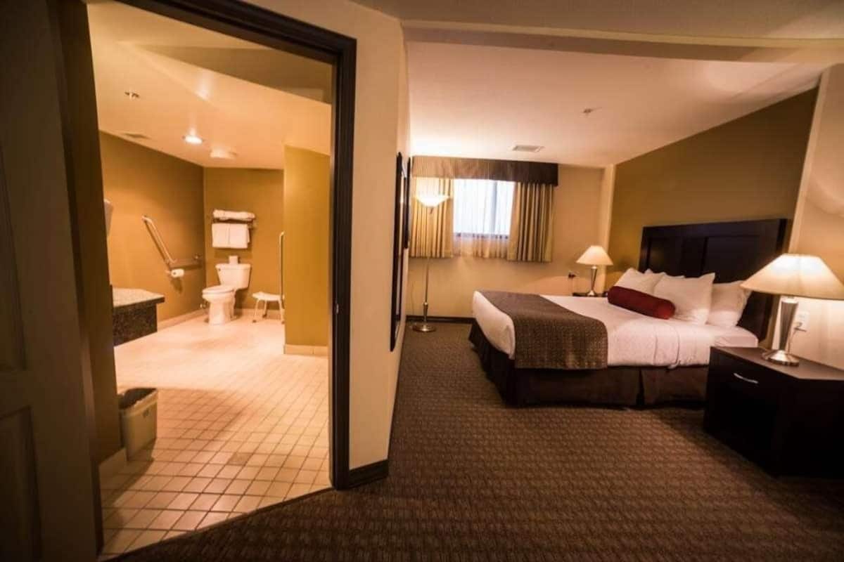 A hotel room with a bed, bedside tables, a lamp, a window with curtains, a bathroom with a toilet, and a shower area.