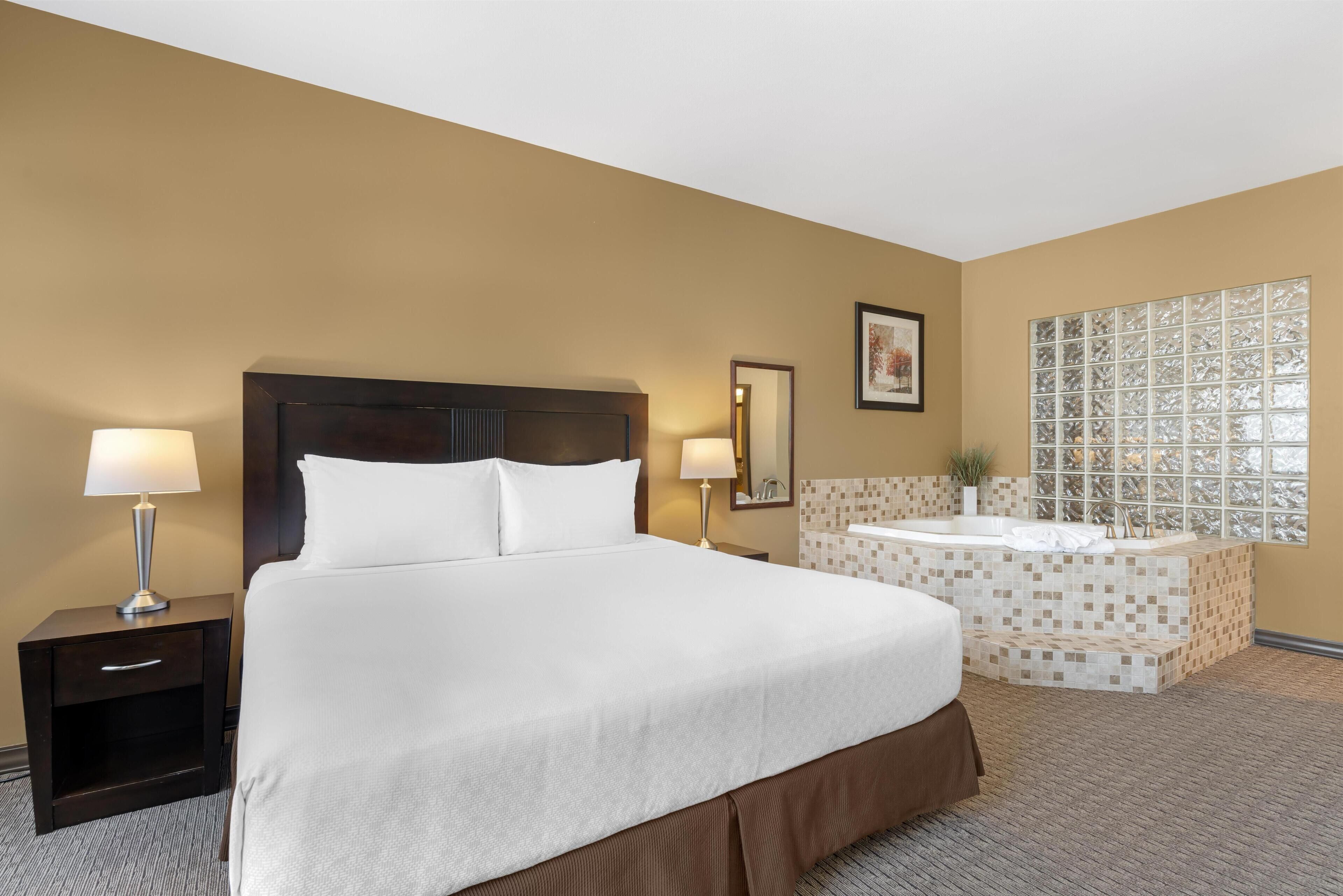 A hotel room with a large bed, a nightstand with a lamp, a bathroom with a bathtub, and a window with a decorative view.