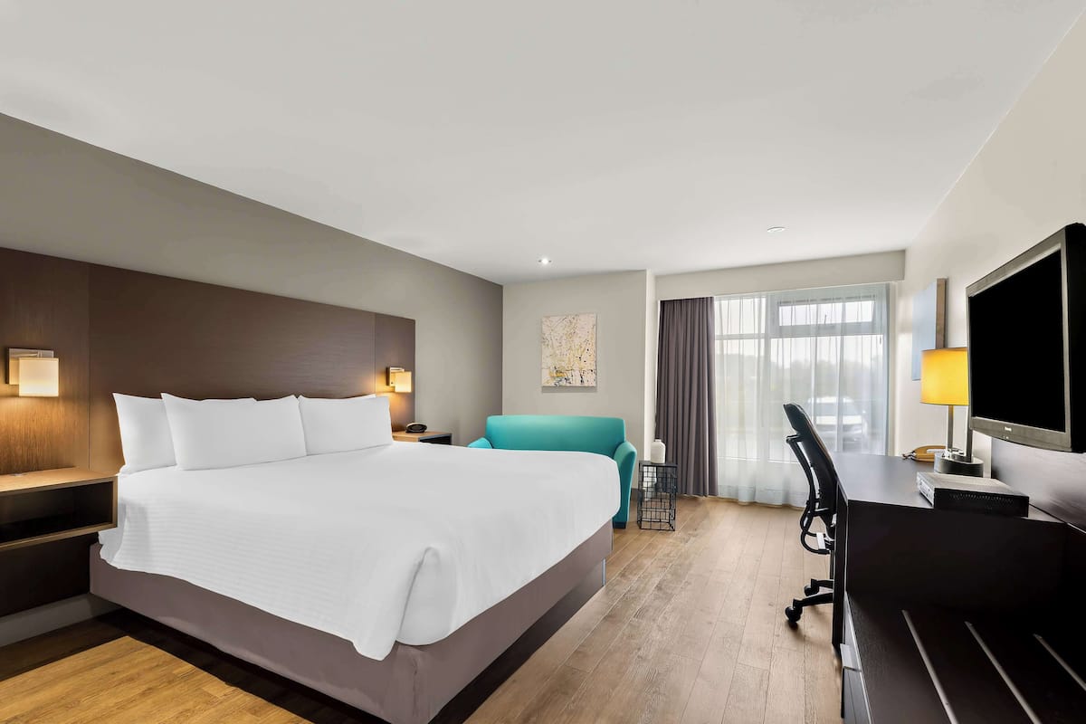Ein Hotelzimmer mit einem großen Bett, einem Schreibtisch mit Computer, einem blaugrünen Sessel und einem Fernseher.