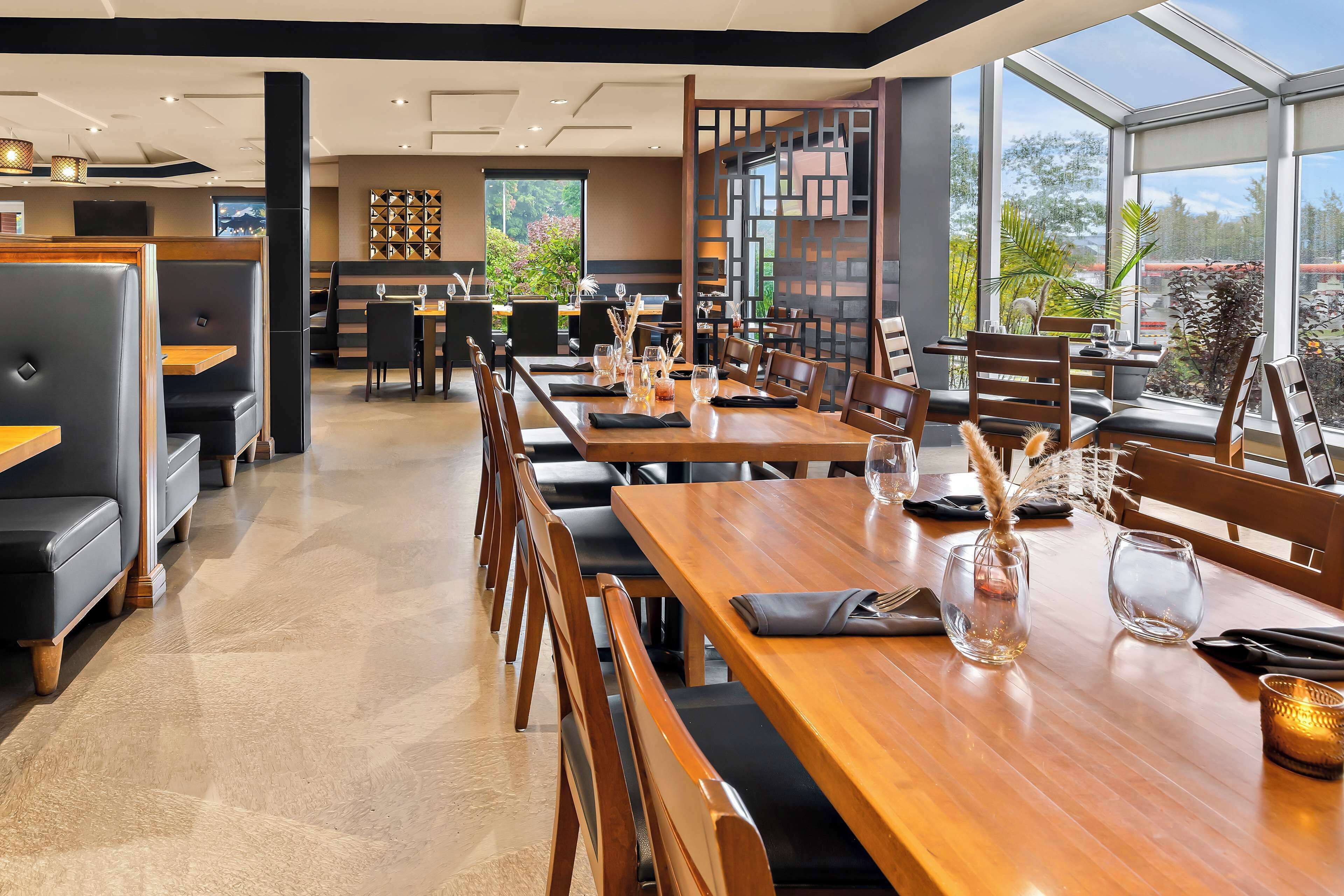 Un restaurante moderno con mesas de madera y cabinas de cuero negro, amplias ventanas con vistas a zonas verdes y un diseño de interiores contemporáneo.
