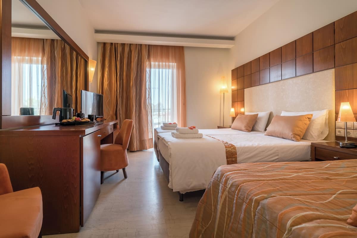 A hotel room with a wooden desk, two beds, a TV, and a large window with curtains.