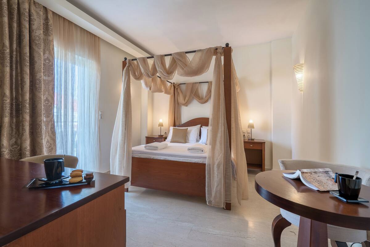 A hotel room with a four-poster bed, a wooden desk, a round table, and a nightstand with a lamp.