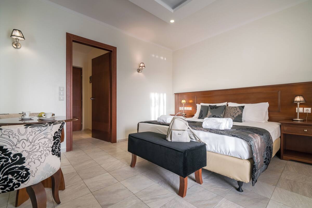 A hotel room with a bed, bedside table, lamp, and a bench with a handbag.
