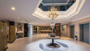 Un hall d’entrée spacieux, agrémenté d’un lustre, d’une table ronde avec un vase, et offrant une vue sur la cuisine et les ascenseurs.
