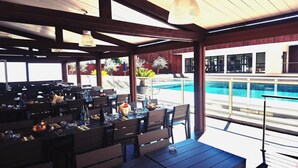 Un espace extérieur aménagé pour les repas, avec des tables et des chaises, une piscine en arrière-plan et une partie couverte agrémentée de guirlandes lumineuses.