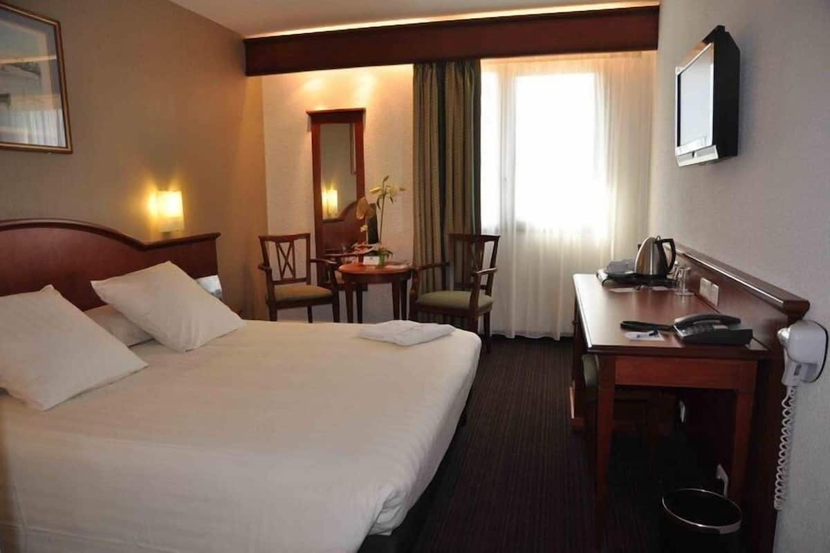 Ein Hotelzimmer mit einem Bett, einem Schreibtisch mit Telefon, einem Sessel, einem kleinen Tisch mit Vase sowie einem an der Wand befestigten Fernseher.