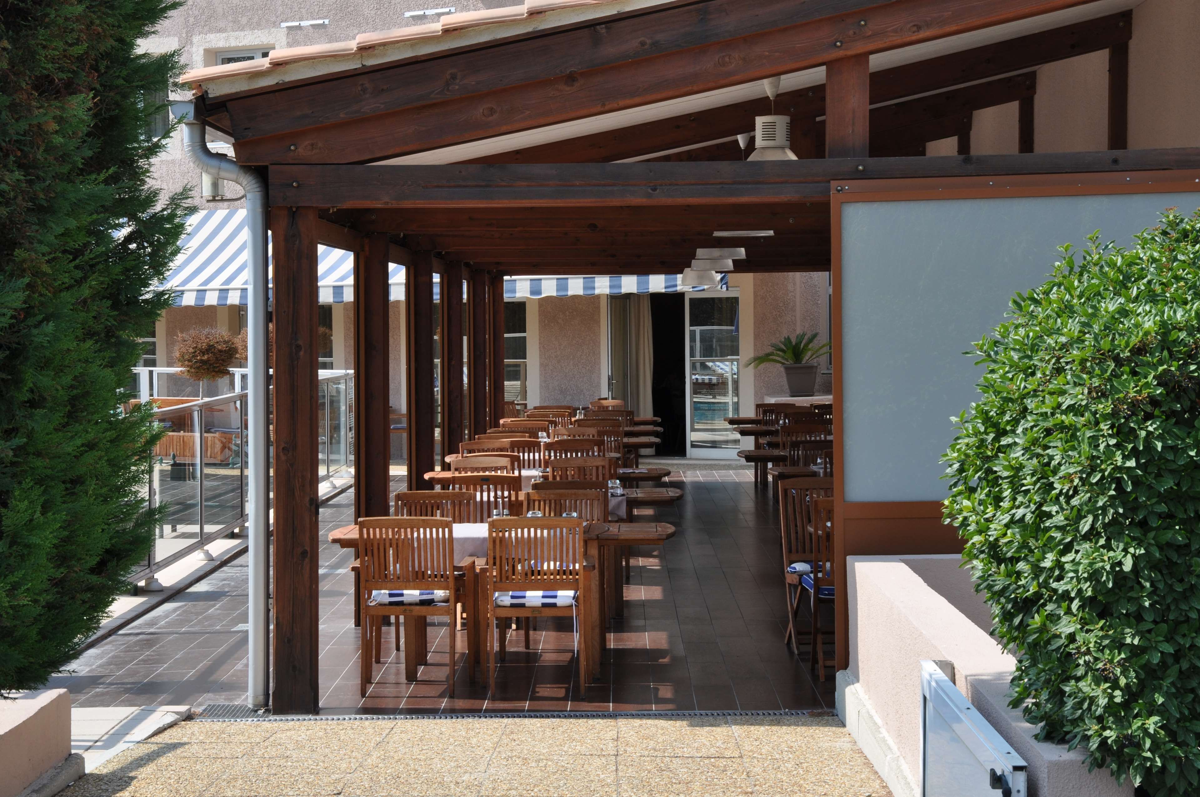 Een overdekt terras met houten tafels en stoelen, een plant in pot, en uitzicht op een gebouw met een gestreept zonnescherm.