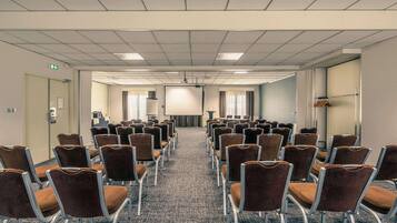 Un salón de conferencias con filas de sillas marrones, piso alfombrado y una pantalla de proyector al frente.