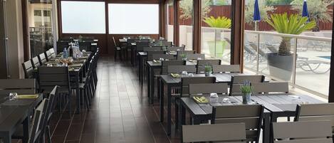 A restaurant with a long counter, multiple tables, and chairs. There are potted plants on the tables, and the restaurant has large windows.