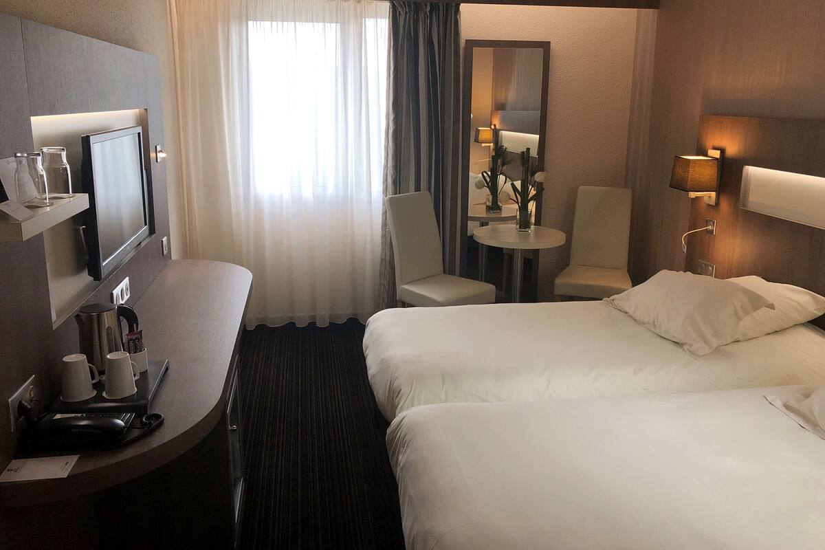 Ein Hotelzimmer mit einem Bett, einem Schreibtisch mit Computer, einem Stuhl, einem kleinen Tisch mit Lampe und einem Spiegel an der Wand.
