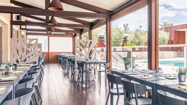 Un restaurant avec de longues tables et des chaises, de grandes fenêtres donnant sur une piscine et un espace verdoyant.