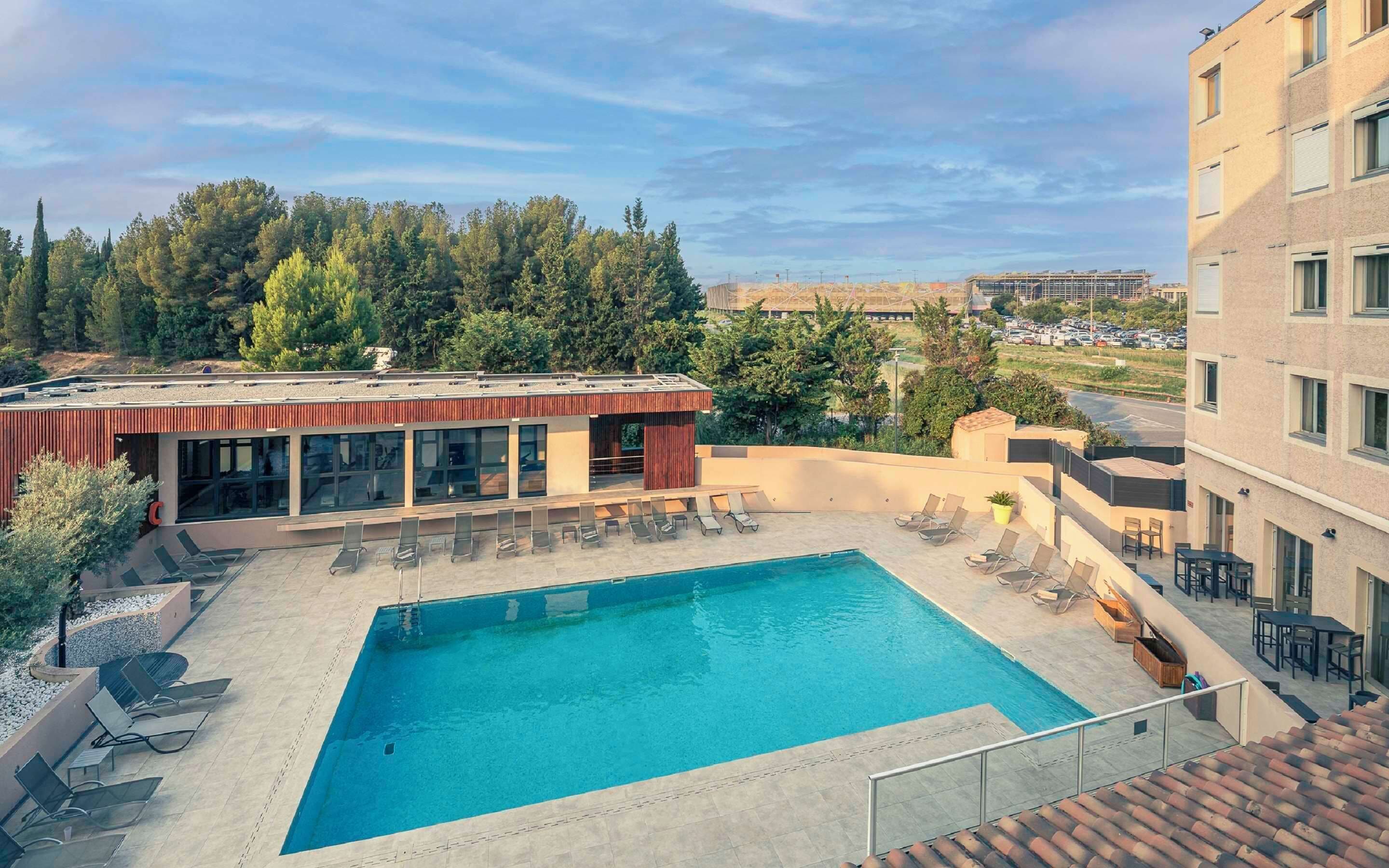 Un espace piscine aménagé sur le toit, avec des chaises longues, une piscine et une vue sur les arbres et les bâtiments en arrière-plan.