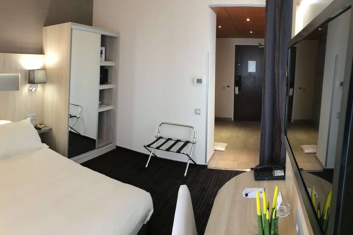 Ein Hotelzimmer mit einem großen Bett, Nachttischen, einem Kleiderschrank, einem Schreibtisch mit Telefon und einem Sessel.