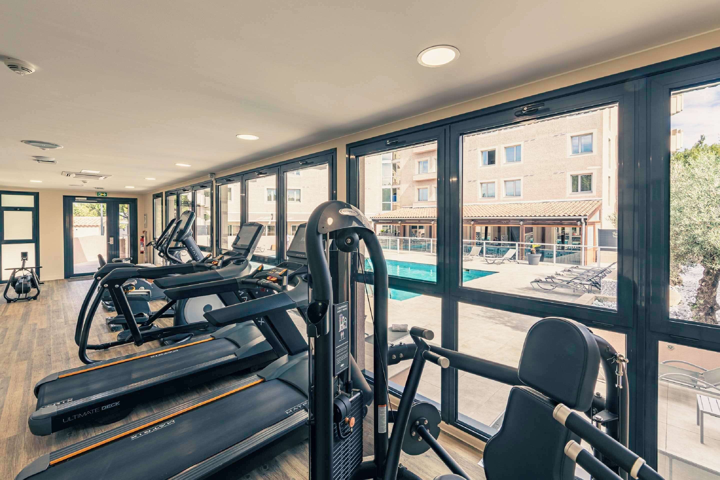 Un gimnasio con cintas de correr, elípticas y pesas sueltas, con amplias ventanas que dan a una piscina y un área de estar al aire libre.
