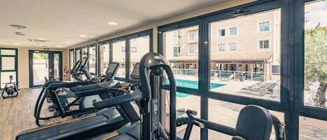 Un gimnasio con cintas de correr, elípticas y pesas sueltas, con amplias ventanas que dan a una piscina y un área de estar al aire libre.
