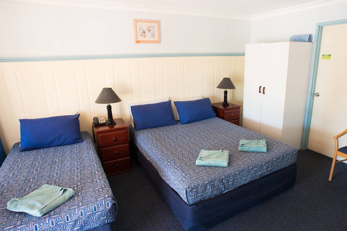 A hotel room with two beds, each with a patterned bedspread and blue pillows, a wooden nightstand with a lamp, a wooden chair, and a wardrobe.