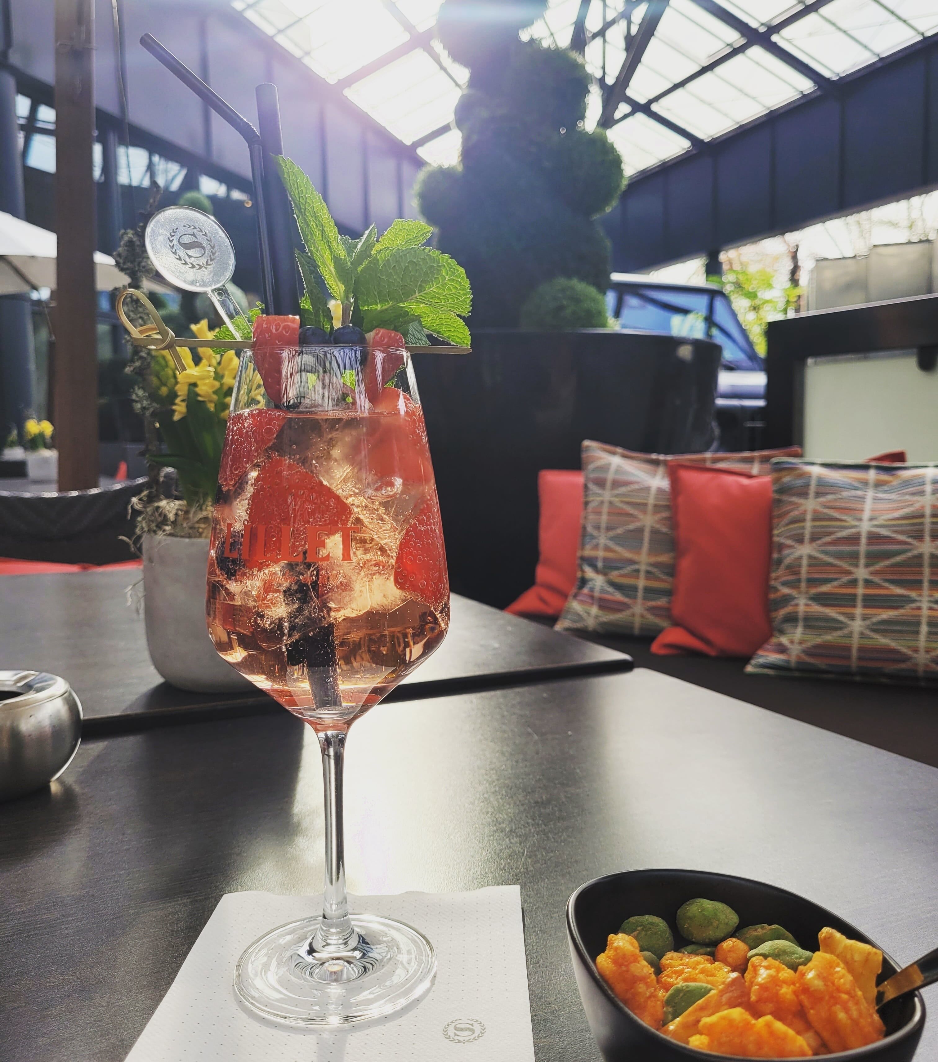 Un vaso de bebida con hielo y fruta sobre una mesa, con una servilleta, un plato de comida y una planta en maceta al fondo.