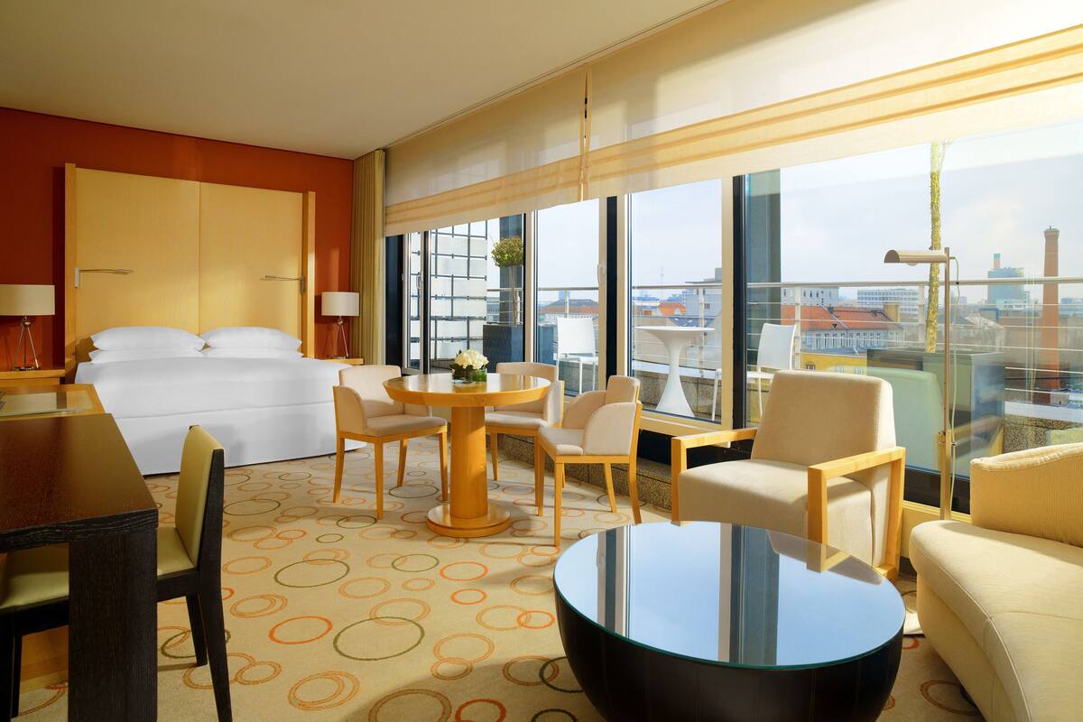 Une chambre d’hôtel avec un lit, une table ronde entourée de chaises, un canapé et de grandes fenêtres donnant sur la ville.