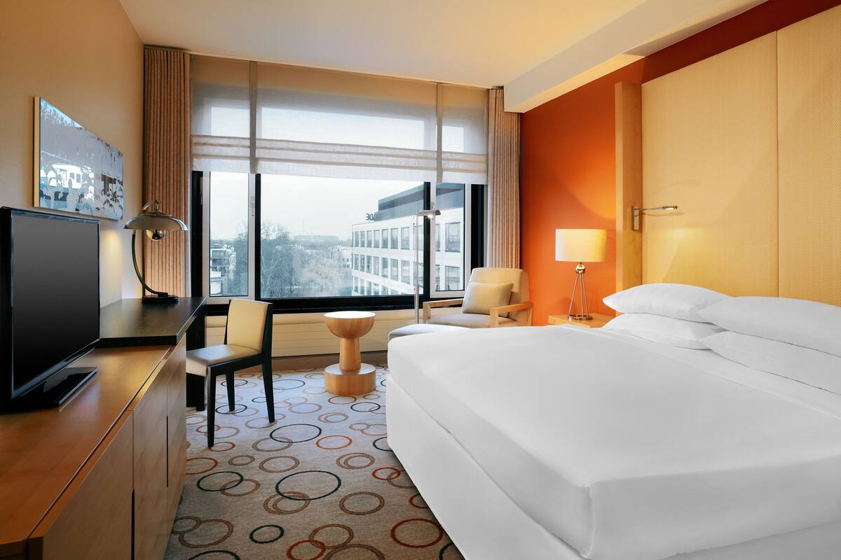 Une chambre d’hôtel avec un lit, un bureau doté d’une télévision, une chaise, une lampe et une vue sur la ville grâce à de grandes fenêtres.