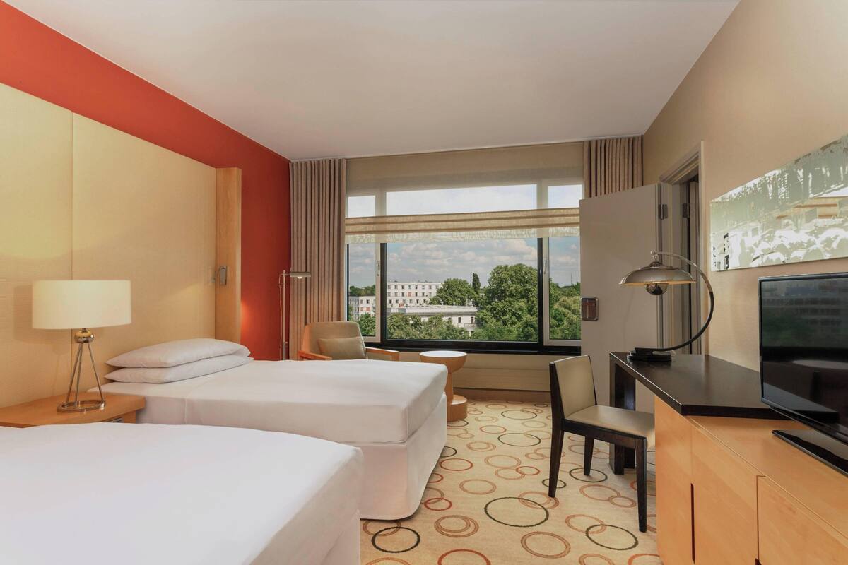 Une chambre d’hôtel avec deux lits, un bureau, une chaise, une télévision et une grande fenêtre donnant sur la ville.