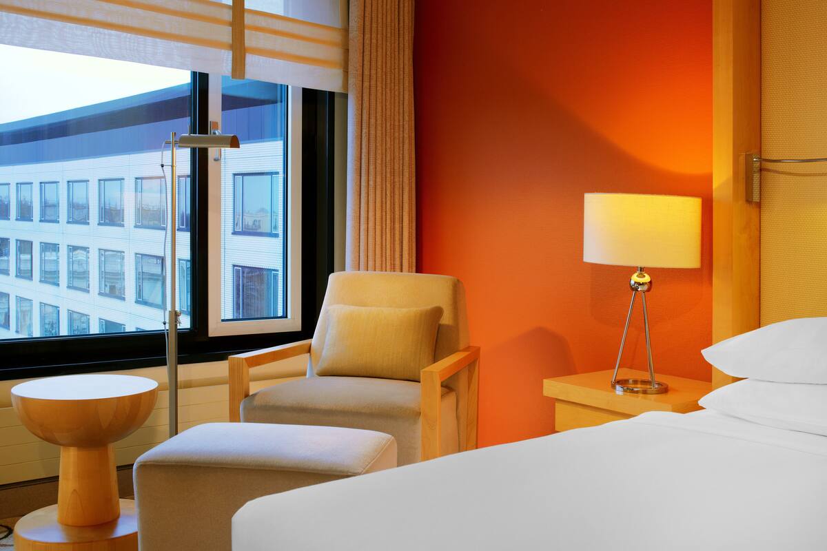 Une chambre d’hôtel avec un lit, deux fauteuils, une petite table, une lampe et une fenêtre donnant sur des immeubles.