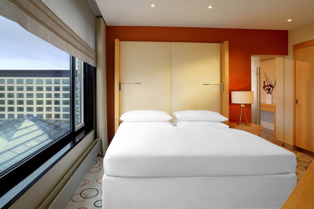 Une chambre d’hôtel avec un grand lit, une fenêtre avec des stores, une vue sur un immeuble et une porte en bois.