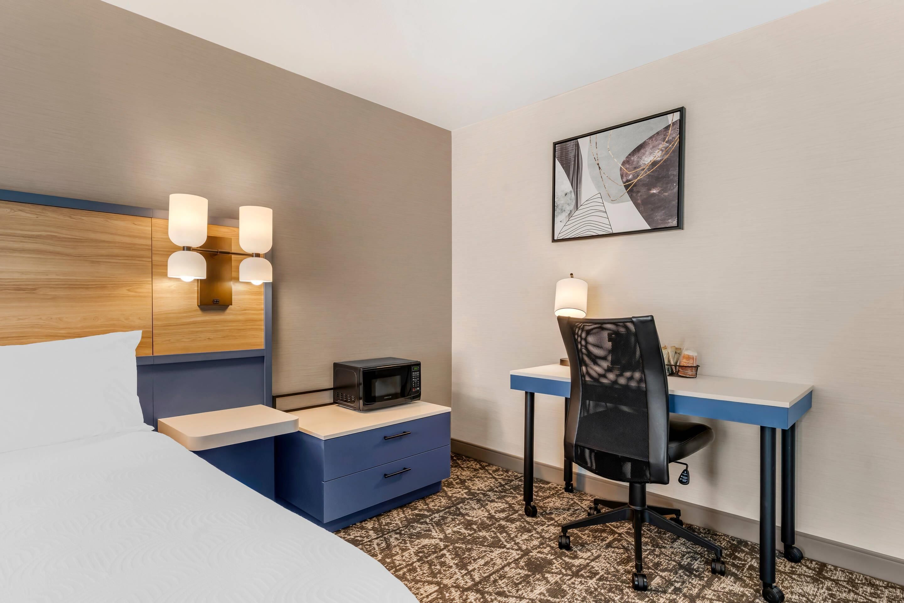 Habitación de hotel con cama, escritorio con silla, microondas y lámpara de pared.