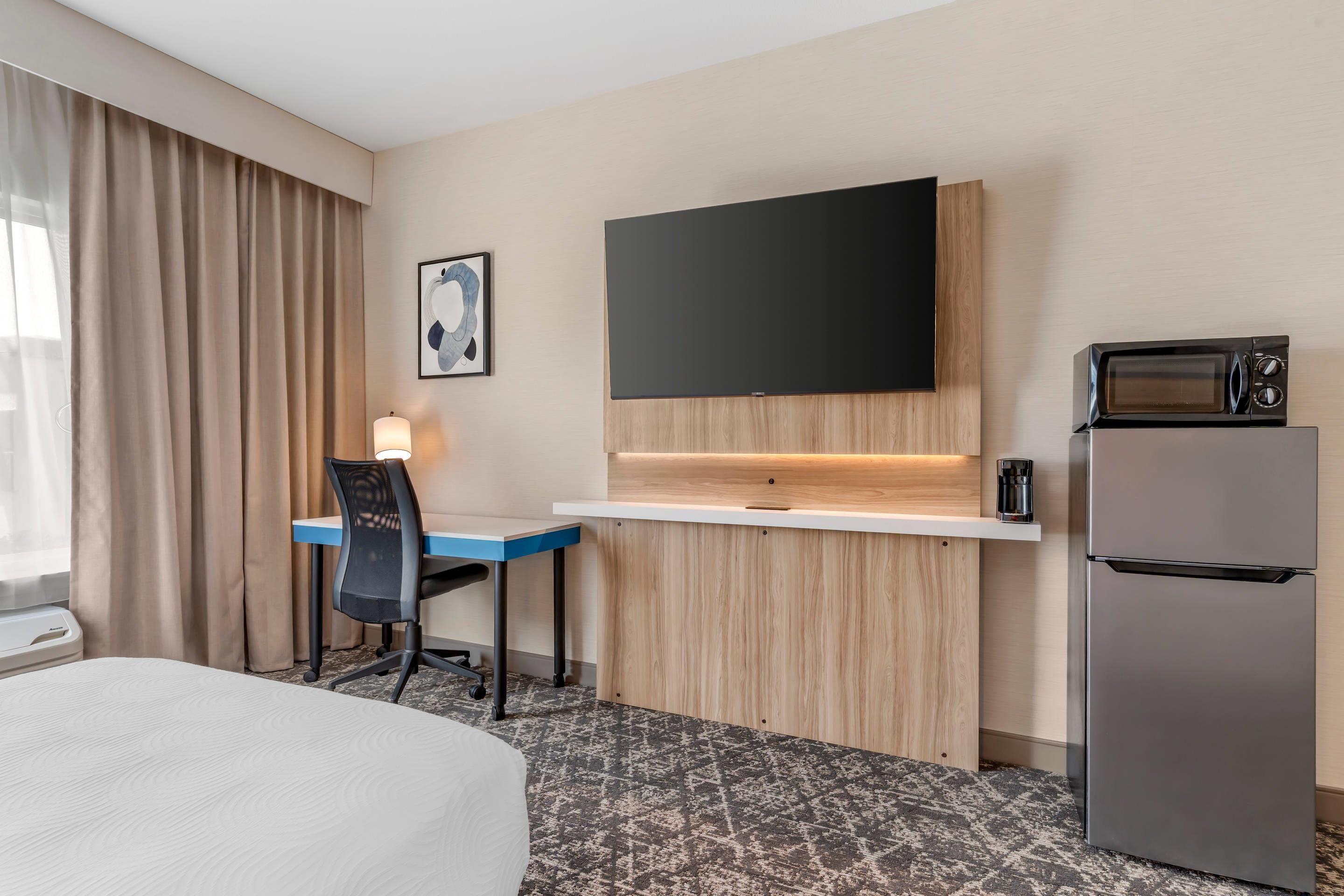 A hotel room with a bed, a desk with a chair, a flat-screen TV mounted on the wall, a microwave, and a refrigerator.