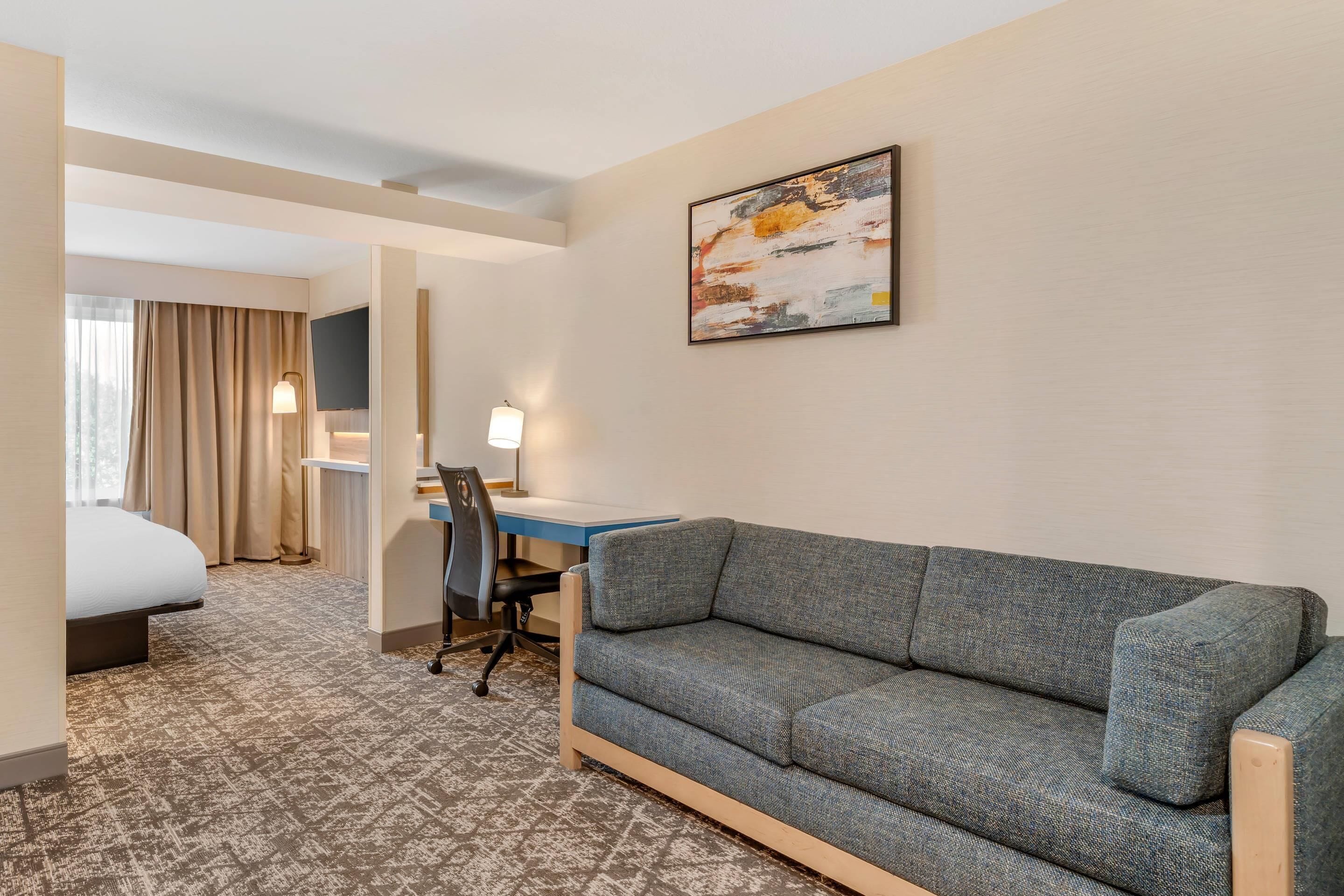 A hotel room with a bed, a desk with a chair, a sofa, a flat-screen TV mounted on the wall, and a patterned carpet.