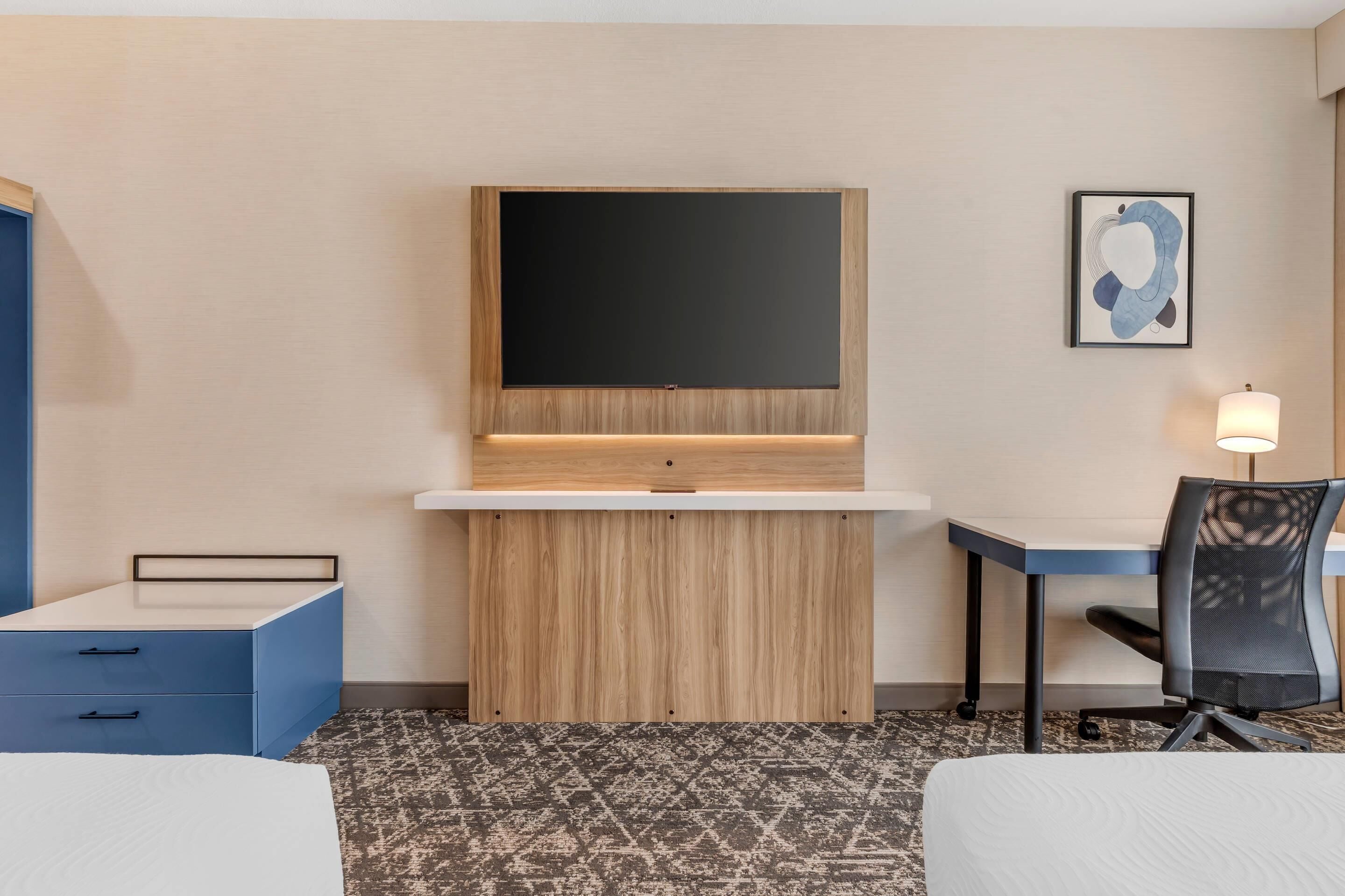 A hotel room with a bed, a desk with a chair, a TV mounted on the wall, and a framed picture.