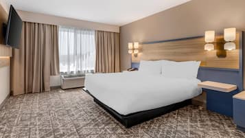 A hotel room with a large bed, a flat-screen TV mounted on the wall, a bedside table with a lamp, and a window with curtains.