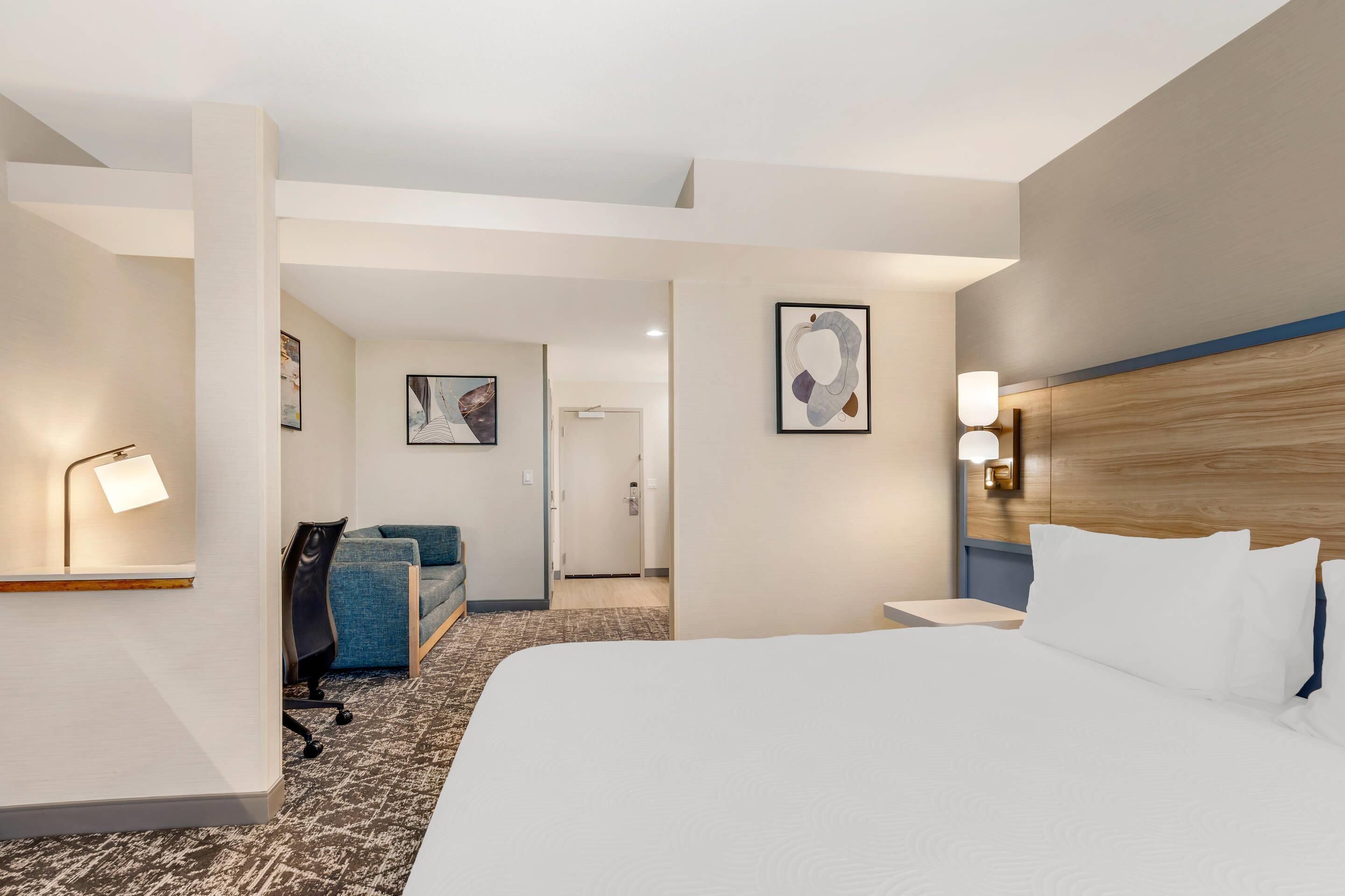 A hotel room with a large bed, a desk with a lamp, a blue chair, and artwork on the walls.