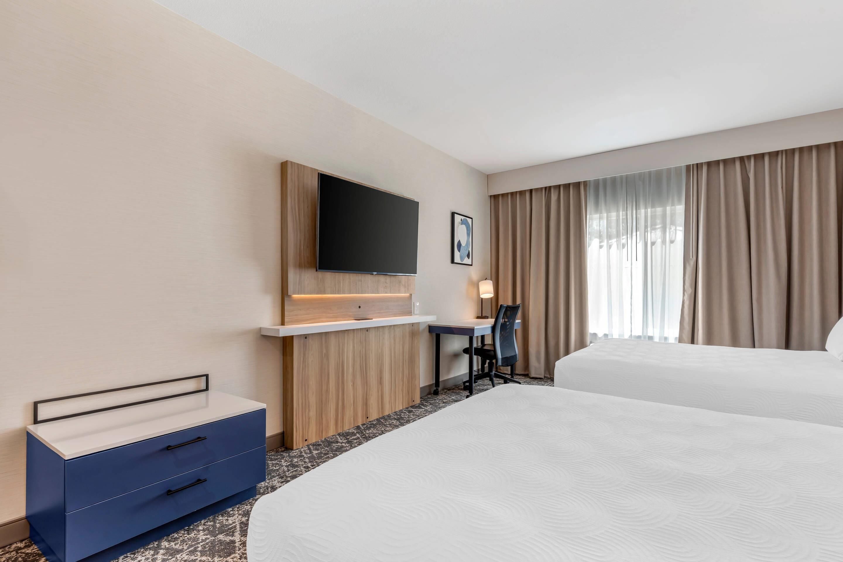 A hotel room with two beds, a desk, a TV mounted on the wall, and a large window with curtains.