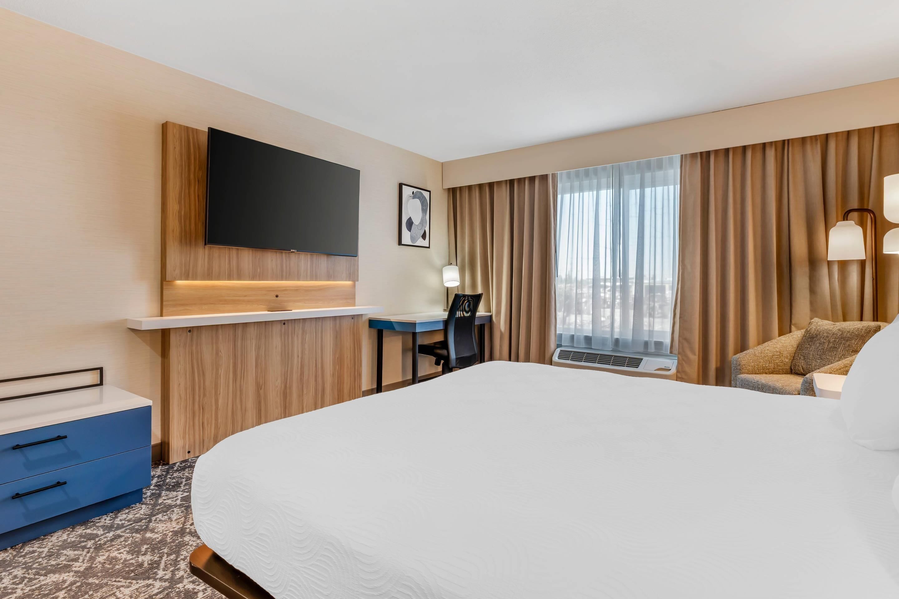 A hotel room with a large bed, a desk, a flat-screen TV, a window with curtains, and a chair.