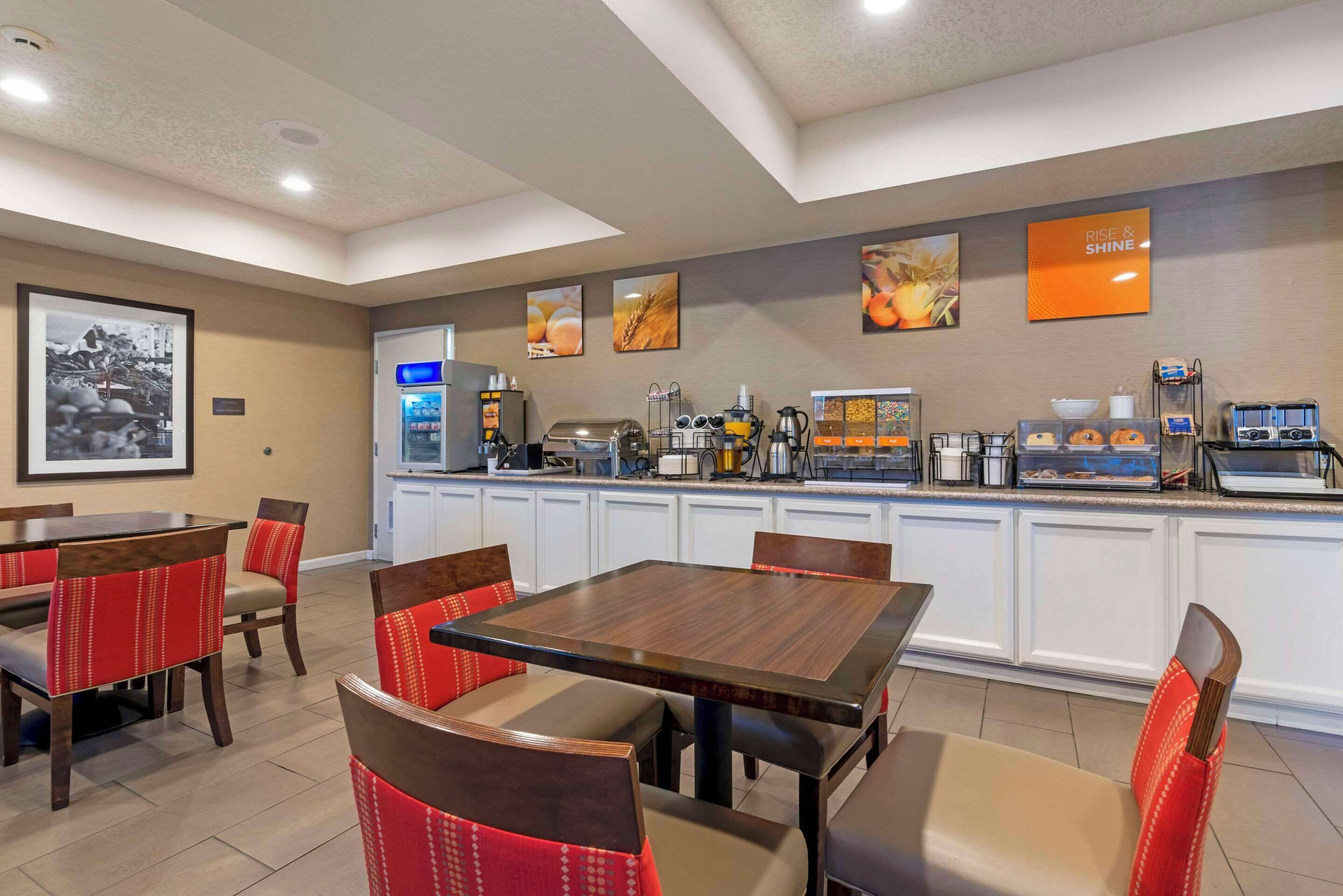 Un rincón para desayunar con una mesa de madera y sillas tapizadas en rojo, una estación de café con distintos electrodomésticos y una obra de arte en la pared que representa alimentos.