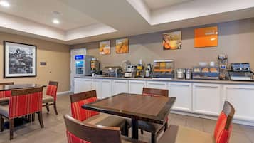 Un rincón para desayunar con una mesa de madera y sillas tapizadas en rojo, una estación de café con distintos electrodomésticos y una obra de arte en la pared que representa alimentos.
