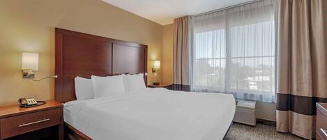 A hotel room with a large bed, a wooden headboard, a bedside table with a lamp, a phone, and a window with curtains.