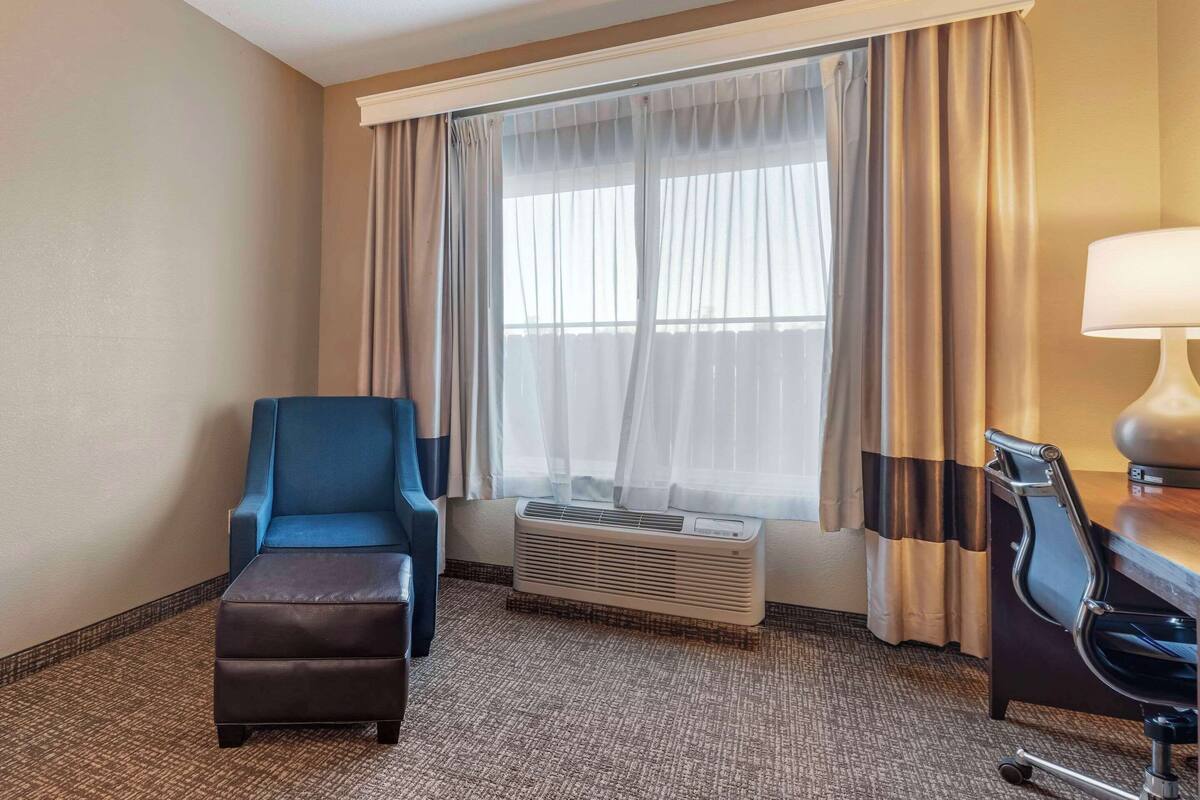 A hotel room with a blue armchair, a brown ottoman, a desk with a lamp, and a large window with sheer curtains.