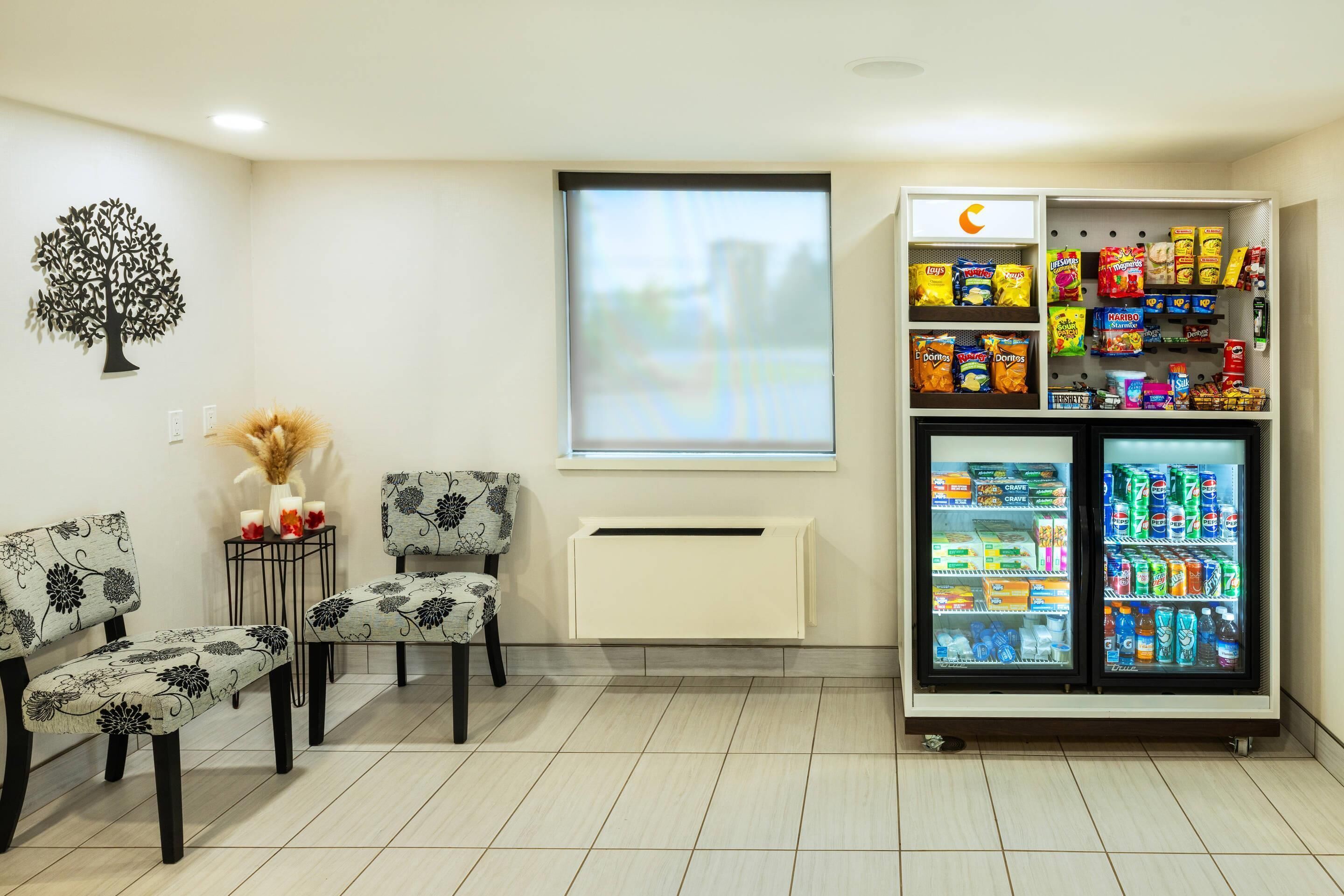 A waiting area with two chairs, a small table with a vase and candles, a wall-mounted tree decoration, a window, and a vending machine stocked with snacks and drinks.