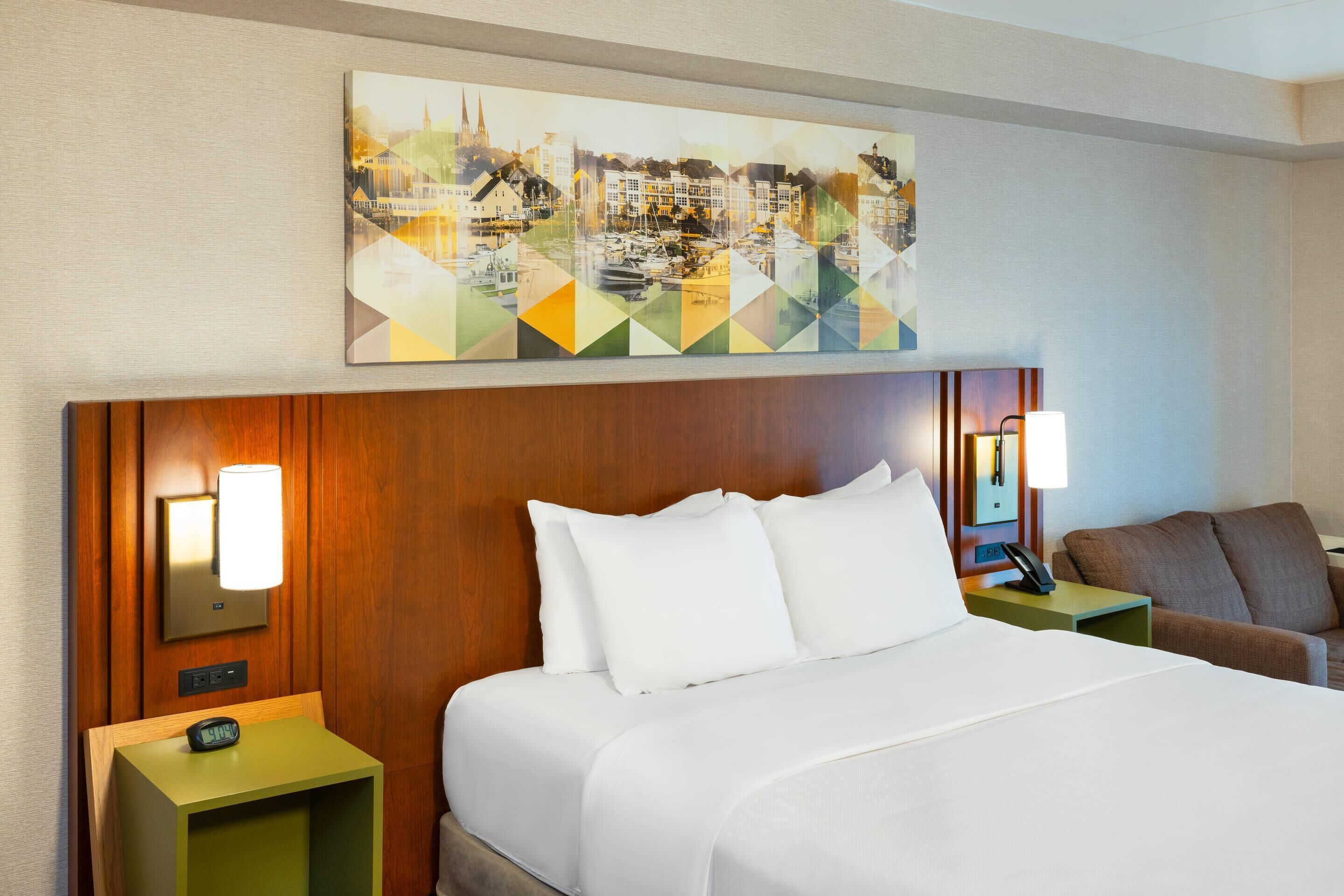 A hotel room with a large bed, two bedside tables with lamps, a wooden headboard, and a cityscape painting above the bed.