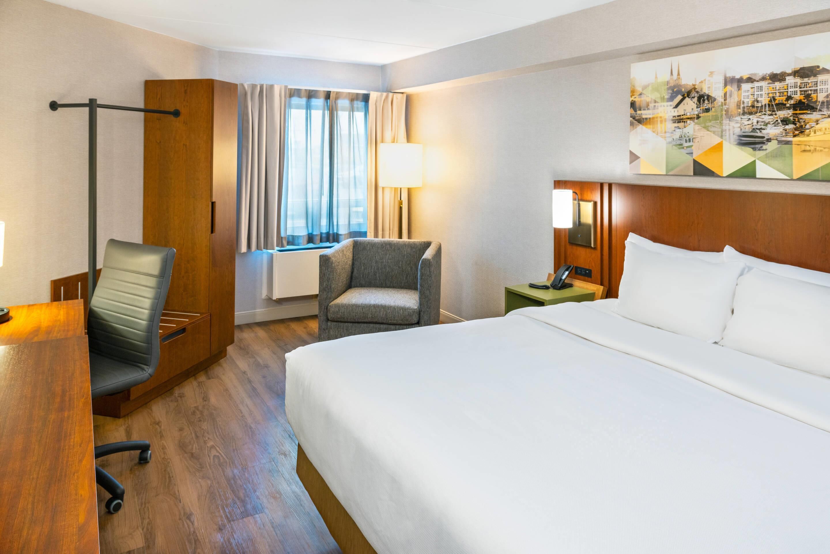 A hotel room with a large bed, a desk, a chair, a window with curtains, a lamp, and a painting on the wall.