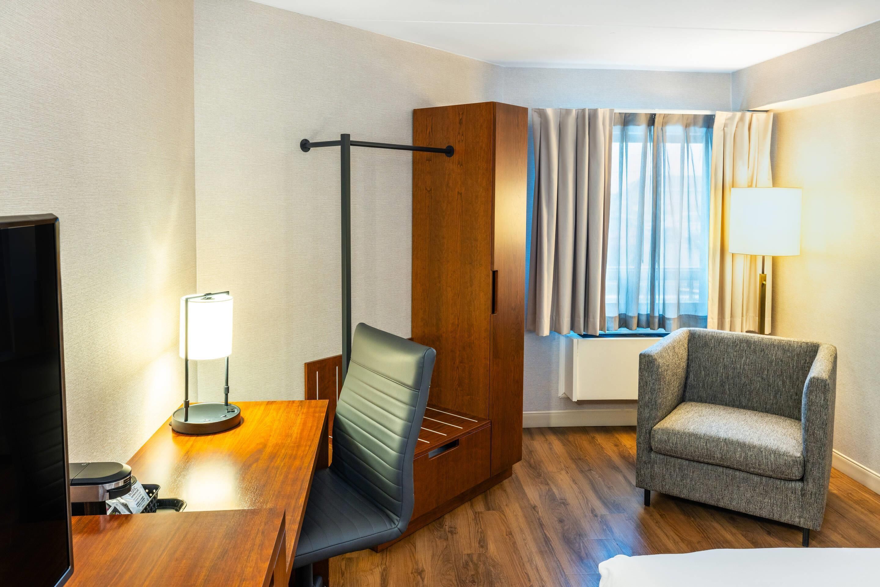 A hotel room with a wooden desk, a chair, a wardrobe, a window with curtains, and a lamp.