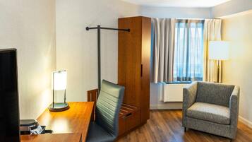 A hotel room with a wooden desk, a chair, a wardrobe, a window with curtains, and a lamp.