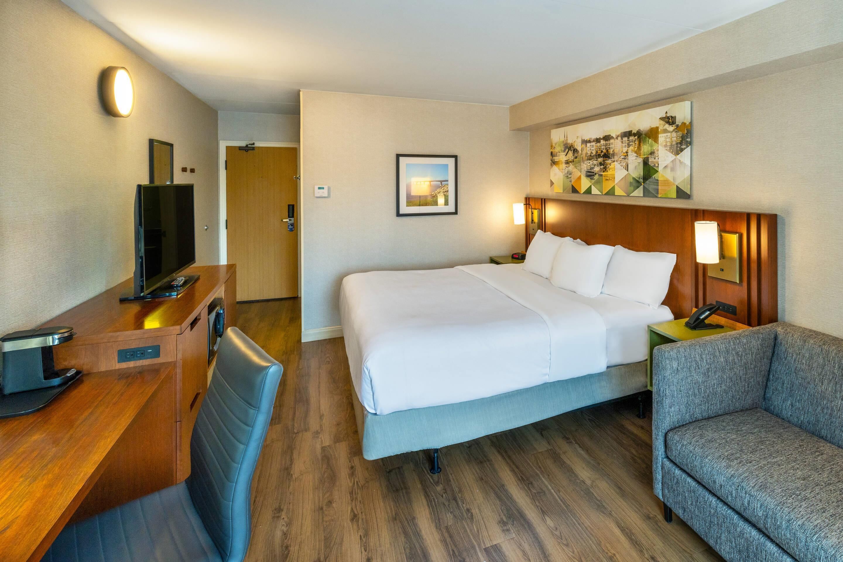A hotel room with a bed, a desk with a computer, a chair, and a painting on the wall.