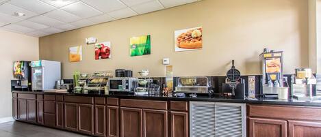 A long counter with various food and drink dispensers, a microwave, and a waffle maker.